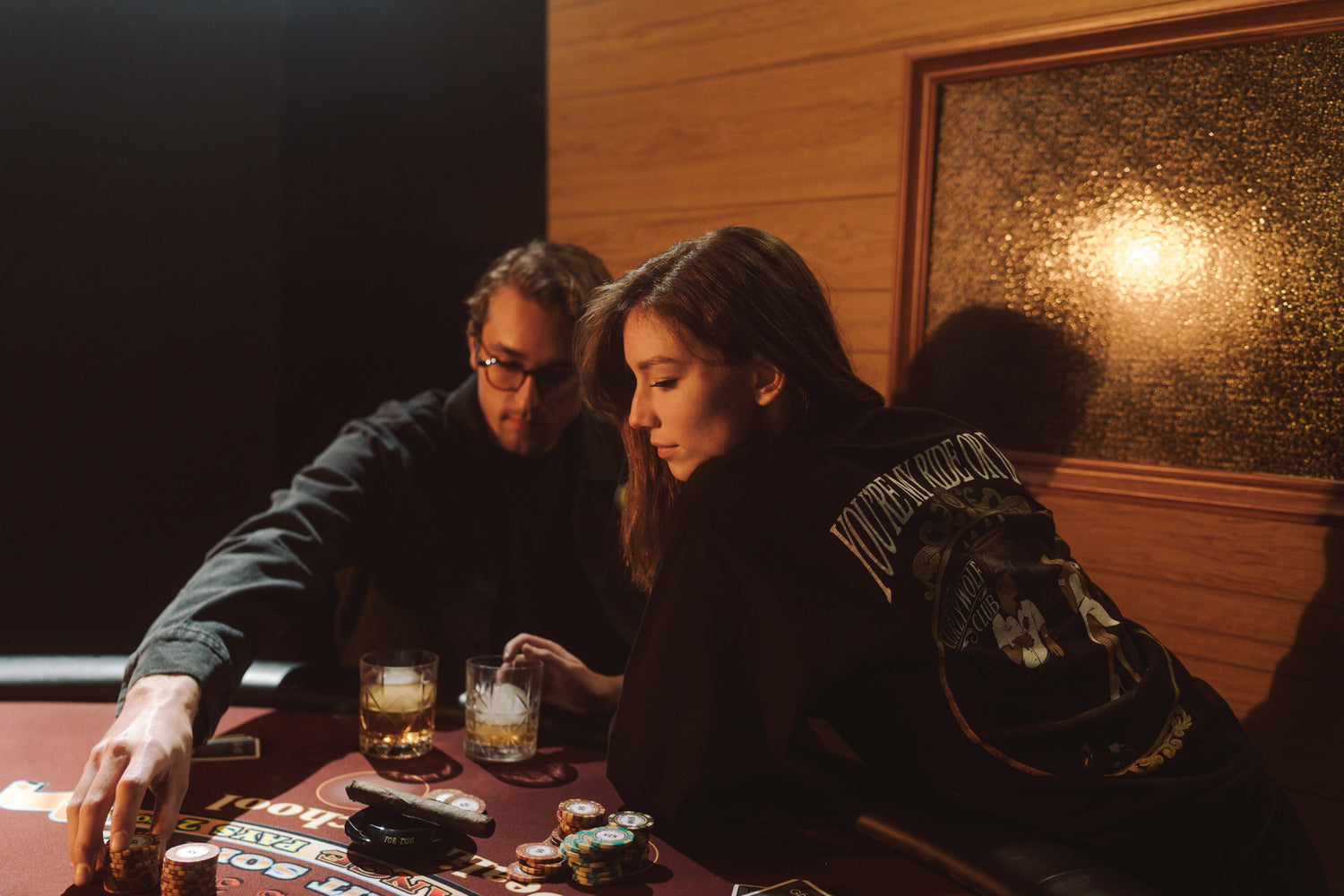 Man and woman sitting in a booth in a dark room playing poker on a poker table and drinking whiskey from two glasses