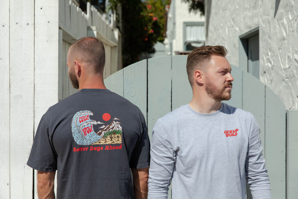Two men in front of a mint green gate wearing the Curly Wolf Maverick tee in Whale Blue and Grey