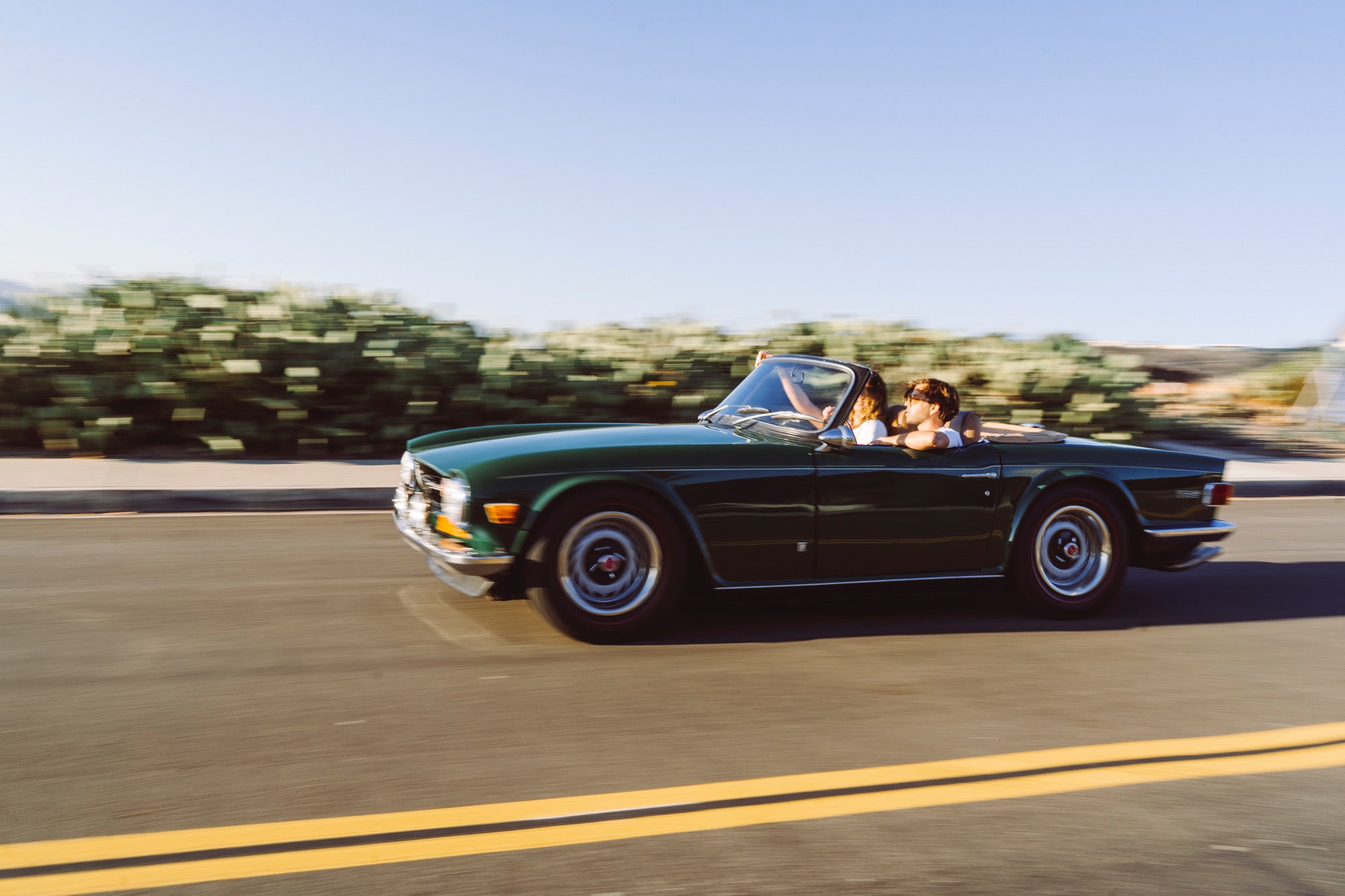 Man and woman driving down the coast in a dark green convertible 