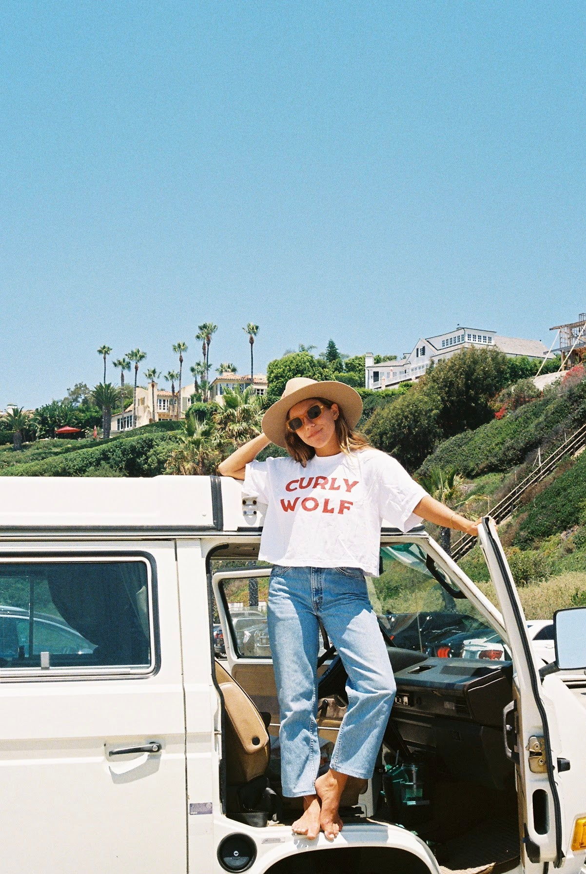Woman standing out of the door of a Volkswagen van wearing a white cropped shirt reading “CURLY WOLF” in red