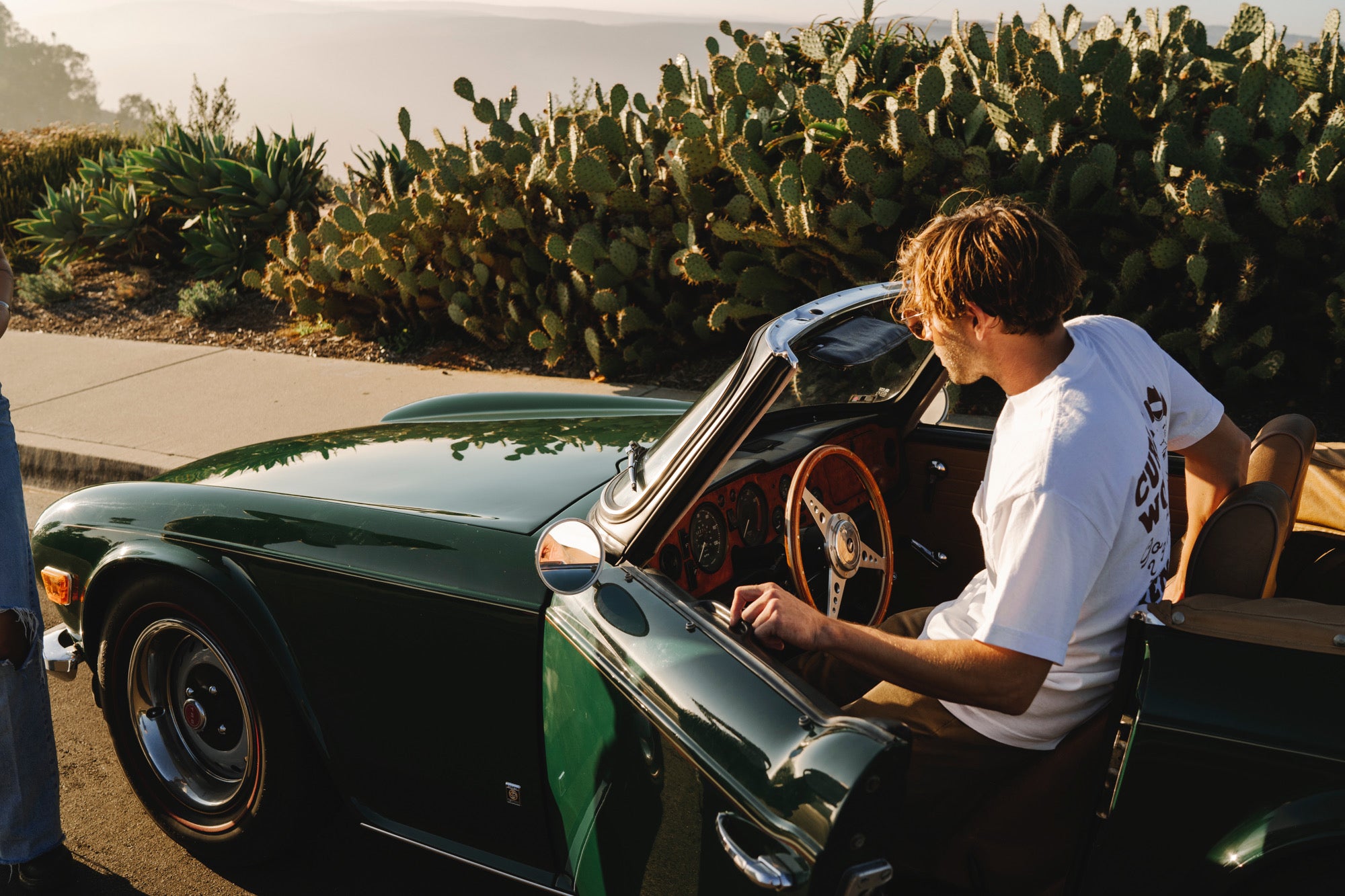 Man stepping into the driver’s seat of a green convertible car wearing a white Curly Wolf shirt. Cacti and ocean cliffs in the background.