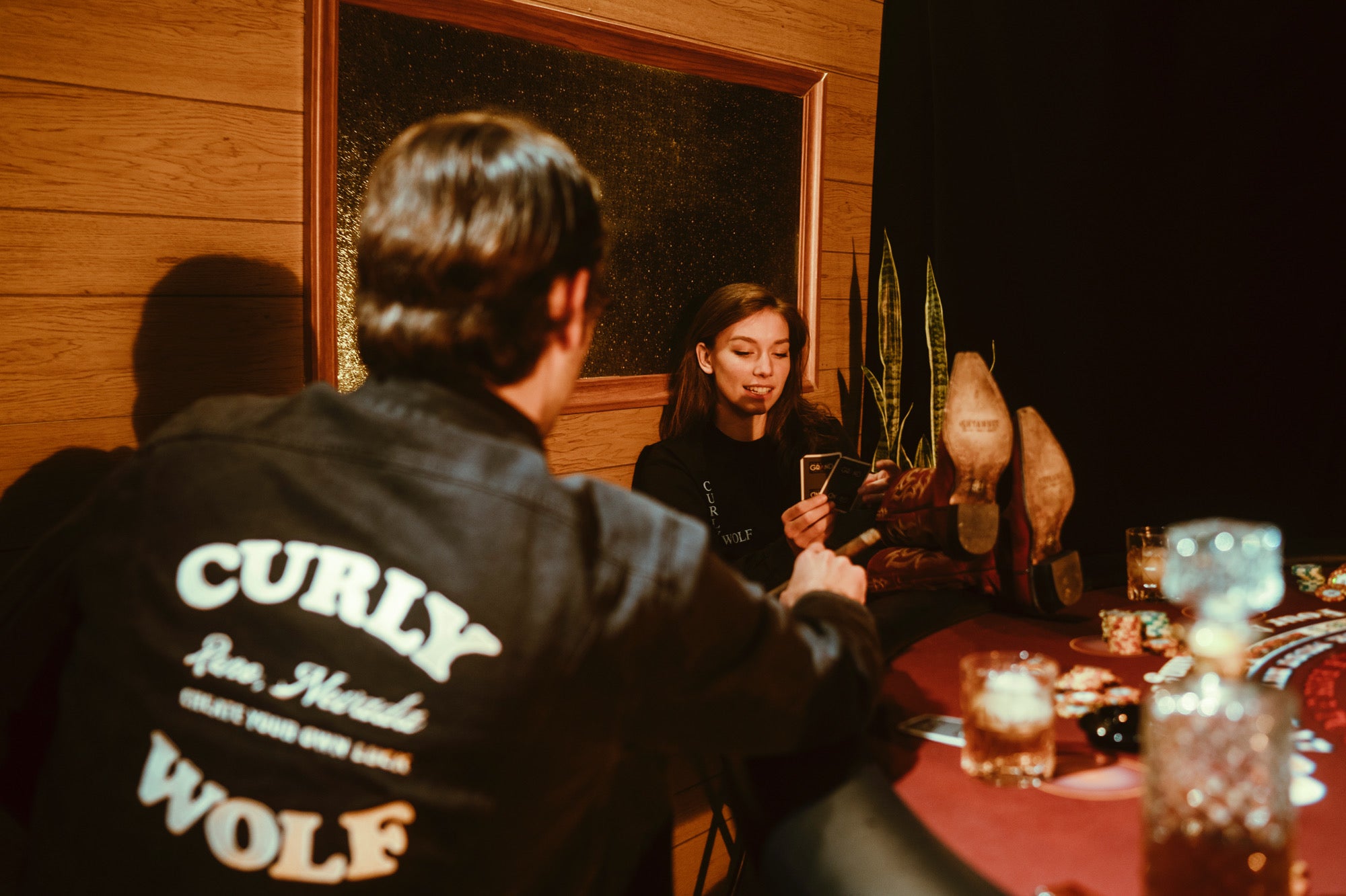 Man and woman playing poker around poker table. Man is wearing Curly Wolf Wells Denim Jacket